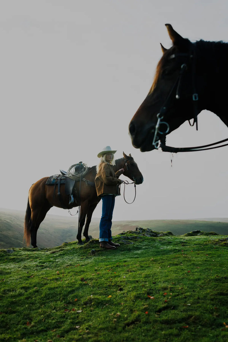 Jenna standing next to a horse on a grassy hill with a misty background and another horses' face in the foreground.