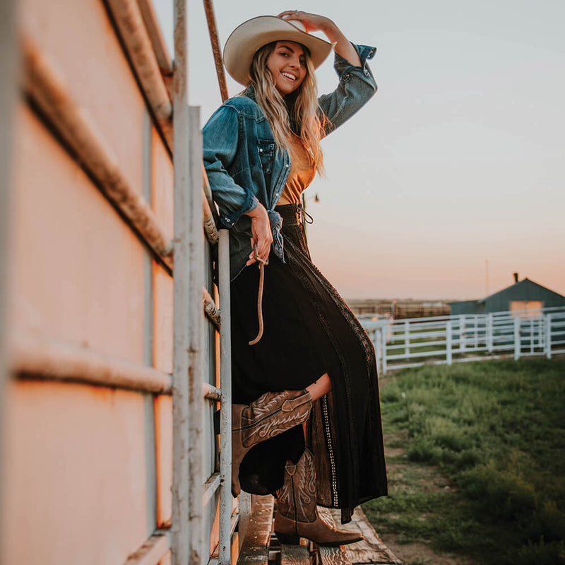 Woman in cowboy hat and Durango boots standing on wooden steps against a sunset sky.