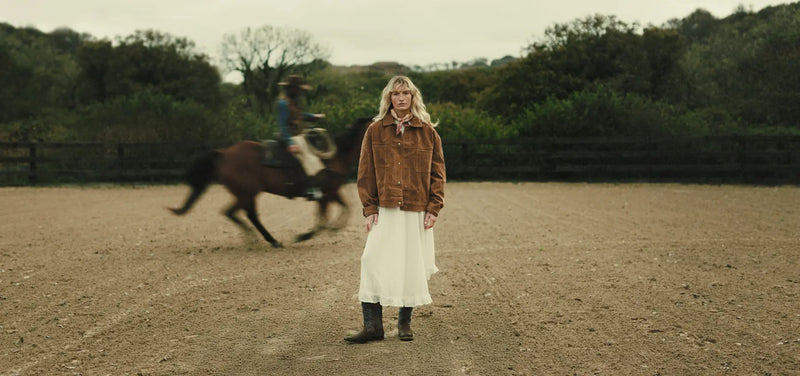 Jenna standing in a dirt field with a horse running in the background.